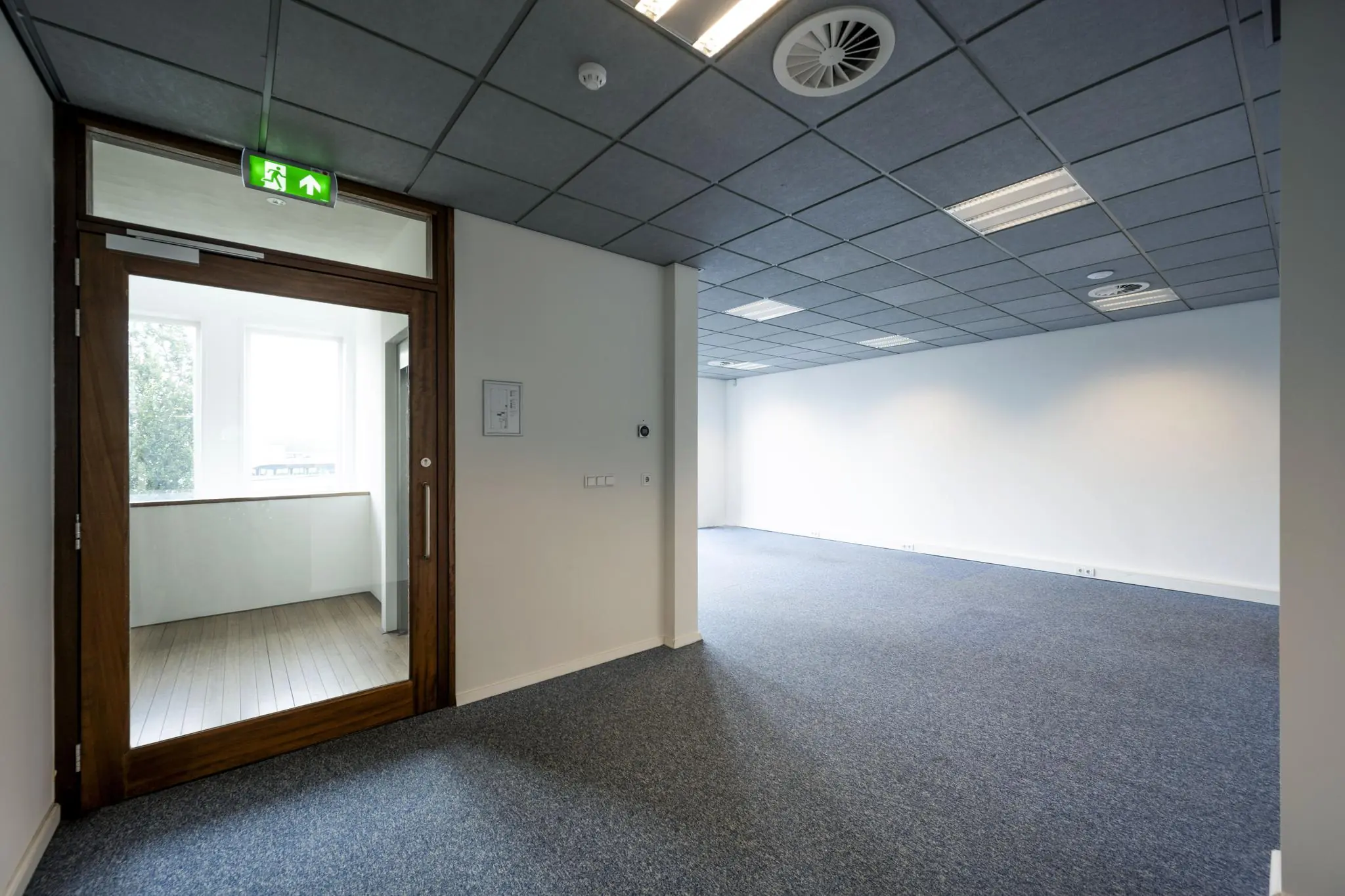 Leeg kantoorruimte aan de De Ruijterkade met systeemplafond, blauwe vloerbedekking en glazen deur met nooduitgangbord.